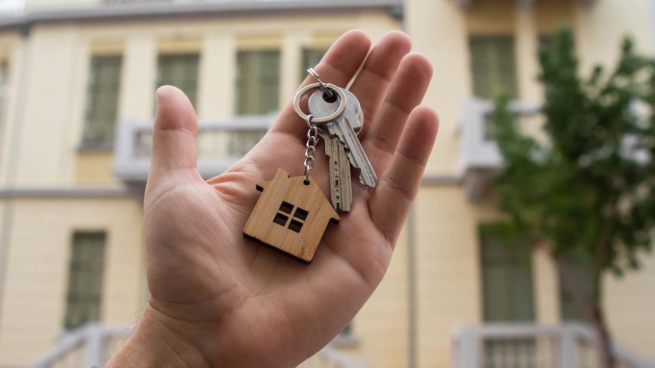 Hand mit Wohnungsschluessel vor Mehrfamilienhaus als Symbol fuer den Kauf einer vermieteten Wohnung und langfristige Kapitalanlage.