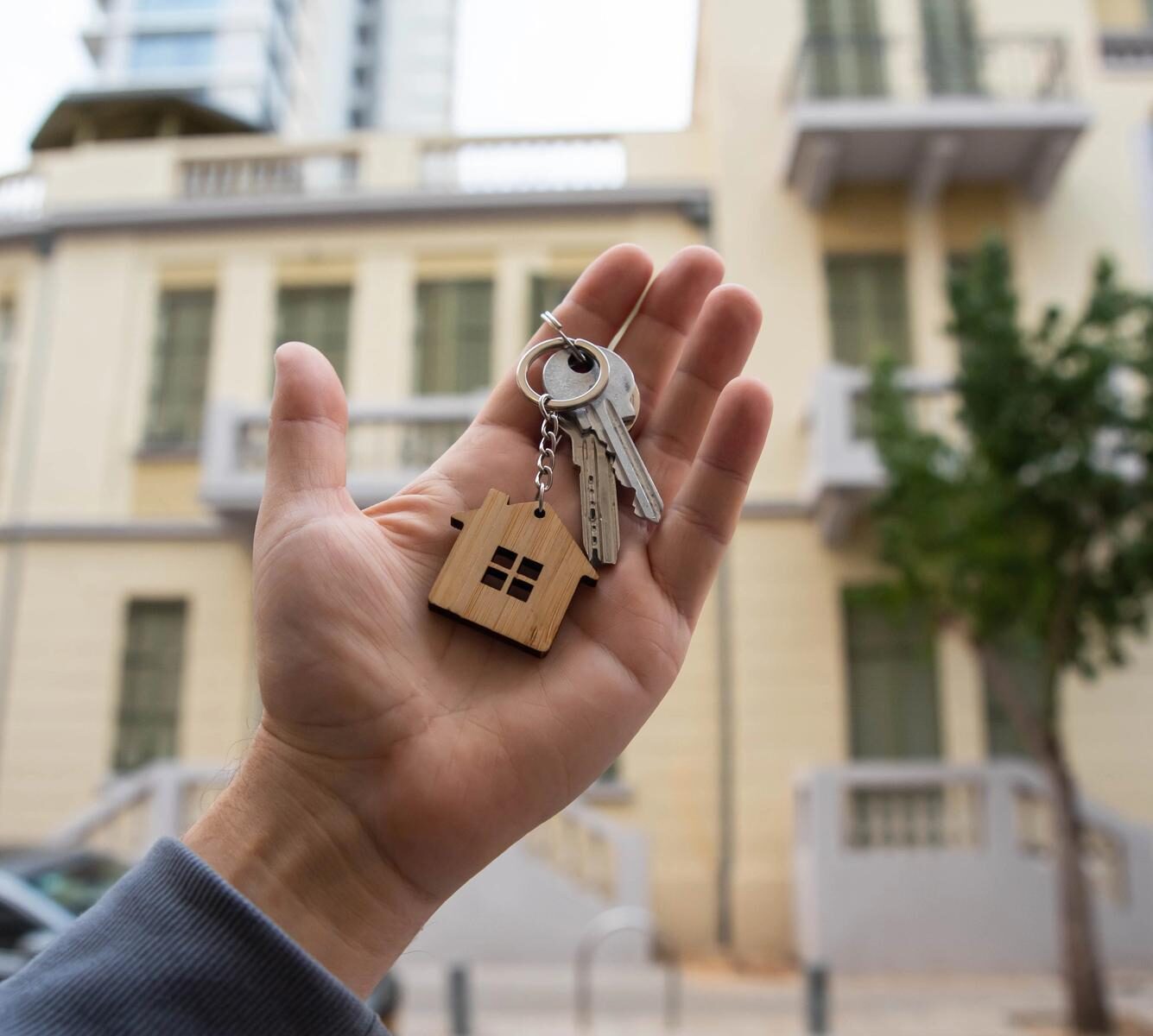 Hand mit Wohnungsschluessel vor Mehrfamilienhaus als Symbol fuer den Kauf einer vermieteten Wohnung und langfristige Kapitalanlage.